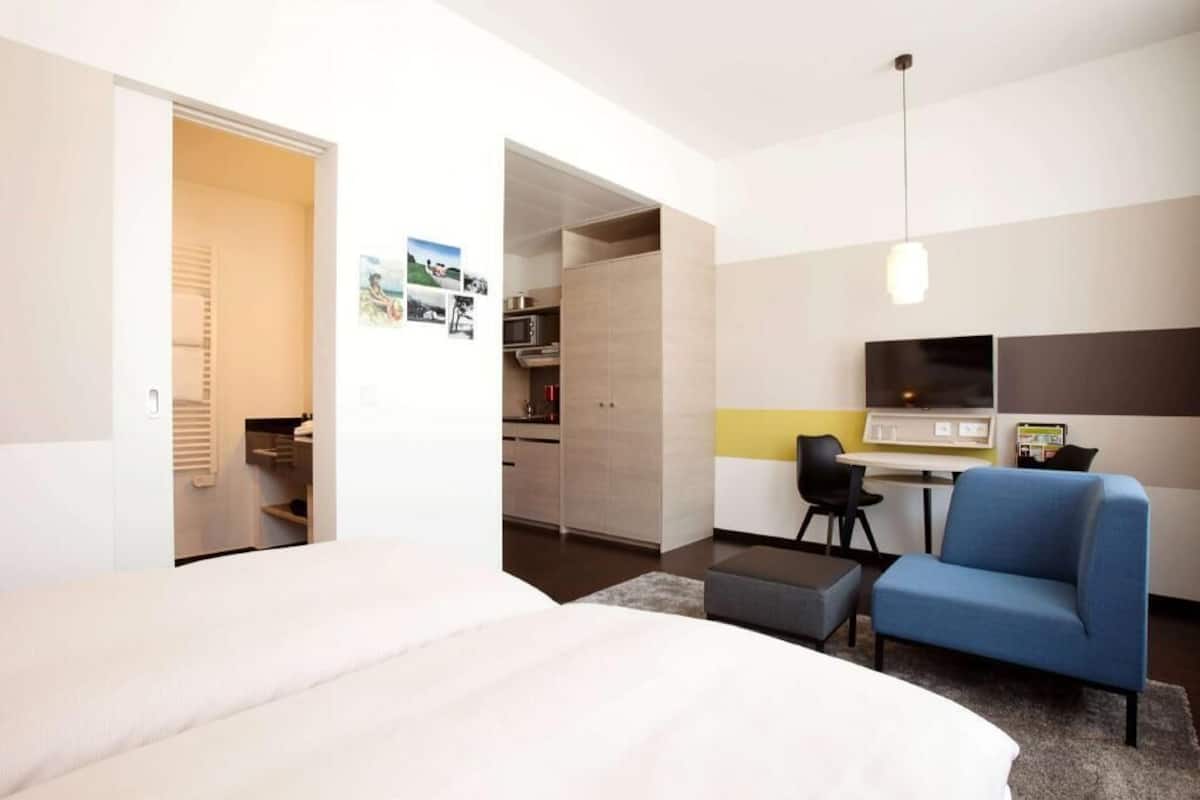 Hotellihuone, jossa on sänky, työpöytä, tuoli ja pieni keittiönurkkaus.