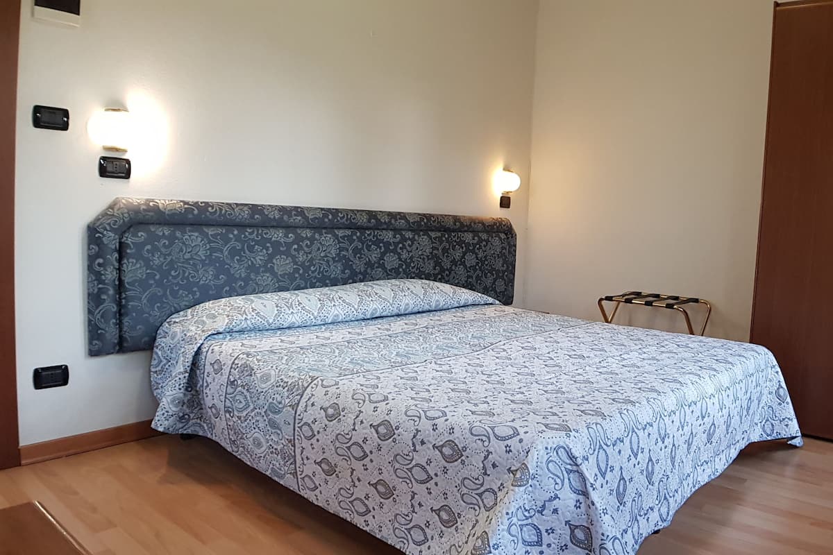 A bedroom with a patterned bedspread, a headboard, bedside lamps, a wooden wardrobe, and a small metal stool.