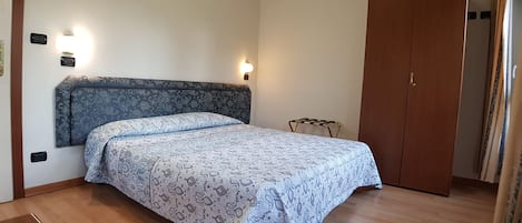 A bedroom with a patterned bedspread, a headboard, bedside lamps, a wooden wardrobe, and a small metal stool.