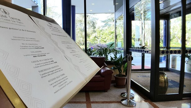 Zona de recepción de hotel con mostrador, cartel de menú y vistas al exterior a través de amplias ventanas.