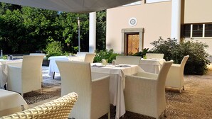 Un área de comedor al aire libre con mesas y sillas de mimbre, cubierta por un toldo blanco, con un fondo de vegetación exuberante y un edificio con puerta y ventana.
