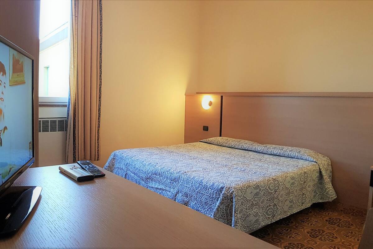 A hotel room with a bed, a desk with a computer and a remote control, a wall-mounted TV, and a bedside lamp.