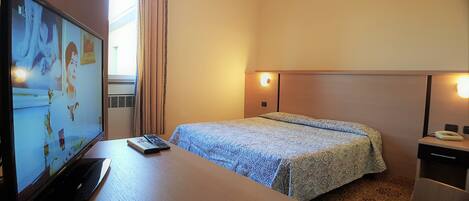 Habitación de hotel con cama, escritorio con computadora y control remoto, televisión montada en la pared y lámpara de noche.