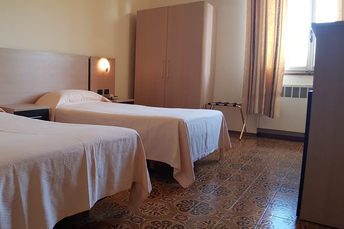A hotel room with two beds, a wardrobe, a nightstand, and a wall-mounted lamp.