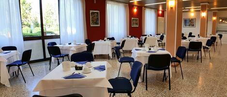 A conference room with multiple tables and chairs, large windows with curtains, and framed pictures on the walls.