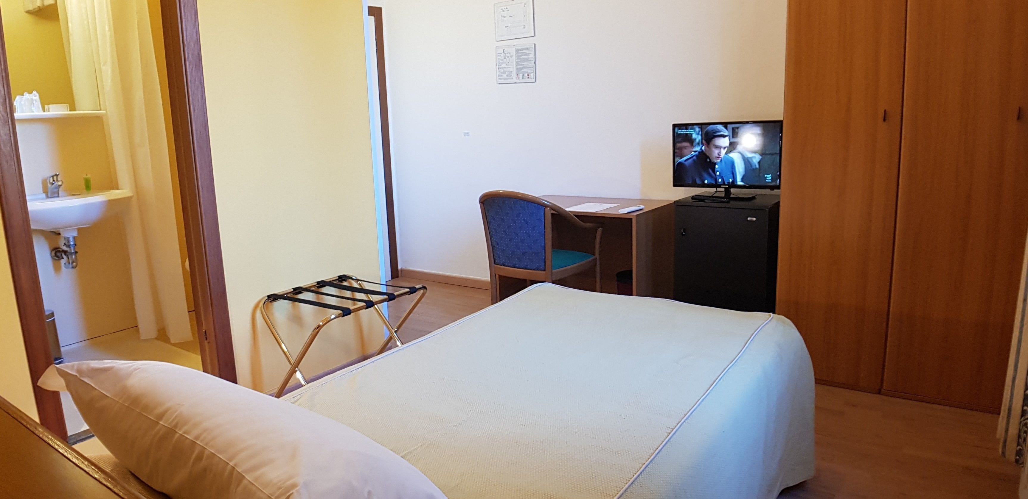 Une chambre d’hôtel comprenant un lit, un bureau avec une chaise, une télévision, un petit placard et une salle de bain avec un lavabo et un miroir.