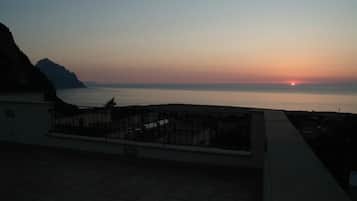 Una terraza en la azotea con vistas a la puesta de sol sobre el mar, una montaña al fondo y un edificio de fachada blanca.