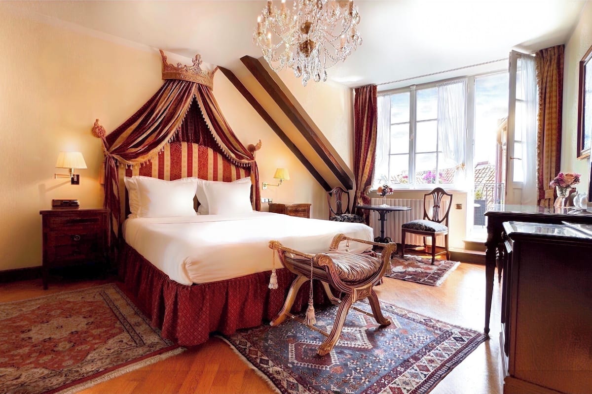 A spacious bedroom with a large bed, a chandelier, a desk, and a view of the outdoors.
