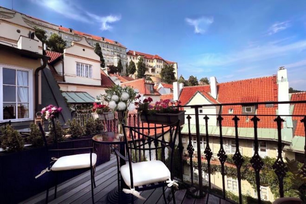 A balcony with a chair and a table, overlooking a cityscape with red-roofed buildings.