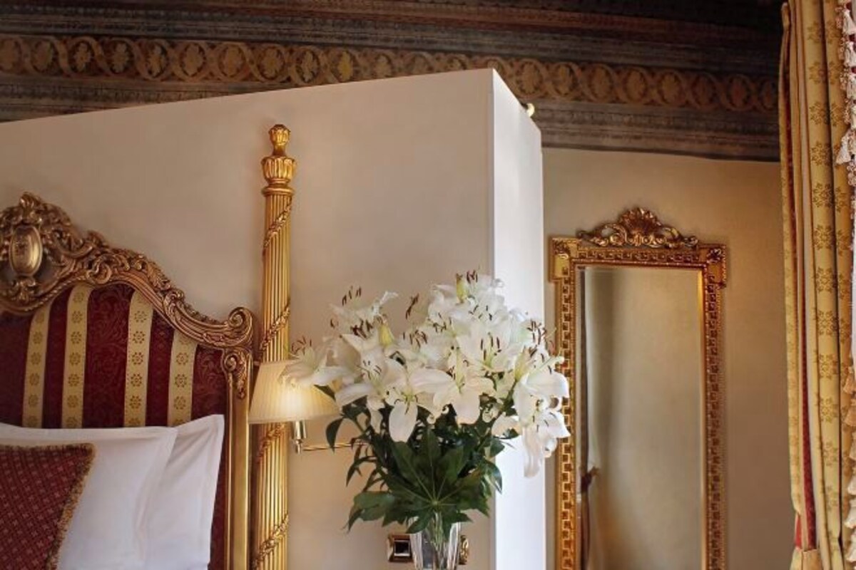 A hotel room with a bed, a vase of white flowers, a wall-mounted lamp, and a mirror with ornate gold detailing.