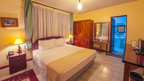 A hotel room with a bed, bedside lamps, a desk with a chair, a mirror, and a bathroom visible through an open door. - The Milimani Lodge (Arusha)