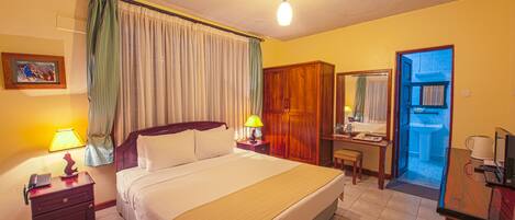 A hotel room with a bed, bedside lamps, a desk with a chair, a mirror, and a bathroom visible through an open door.