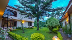 A two-story building with a spiral staircase, a well-maintained garden with various plants and a pathway, and a large tree in the center. - The Milimani Lodge (Arusha)