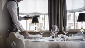 A well-set dining table with multiple wine glasses and cutlery, a waiter in a white shirt and vest, and a window with curtains in the background. - Post Hotel - Adults Only (San Candido)