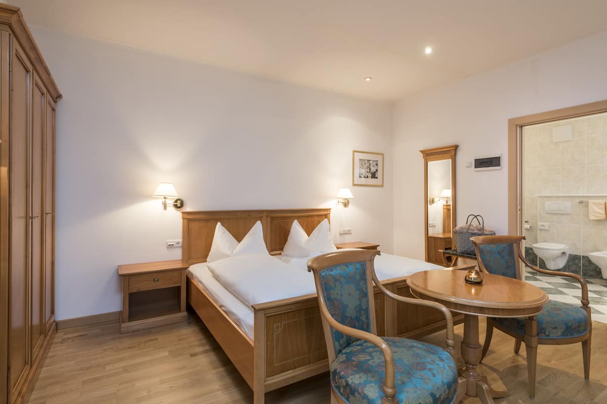 Une chambre d’hôtel avec un lit en bois, deux tables de chevet, une chaise, une table, un miroir et une salle de bain visible à travers une porte ouverte.