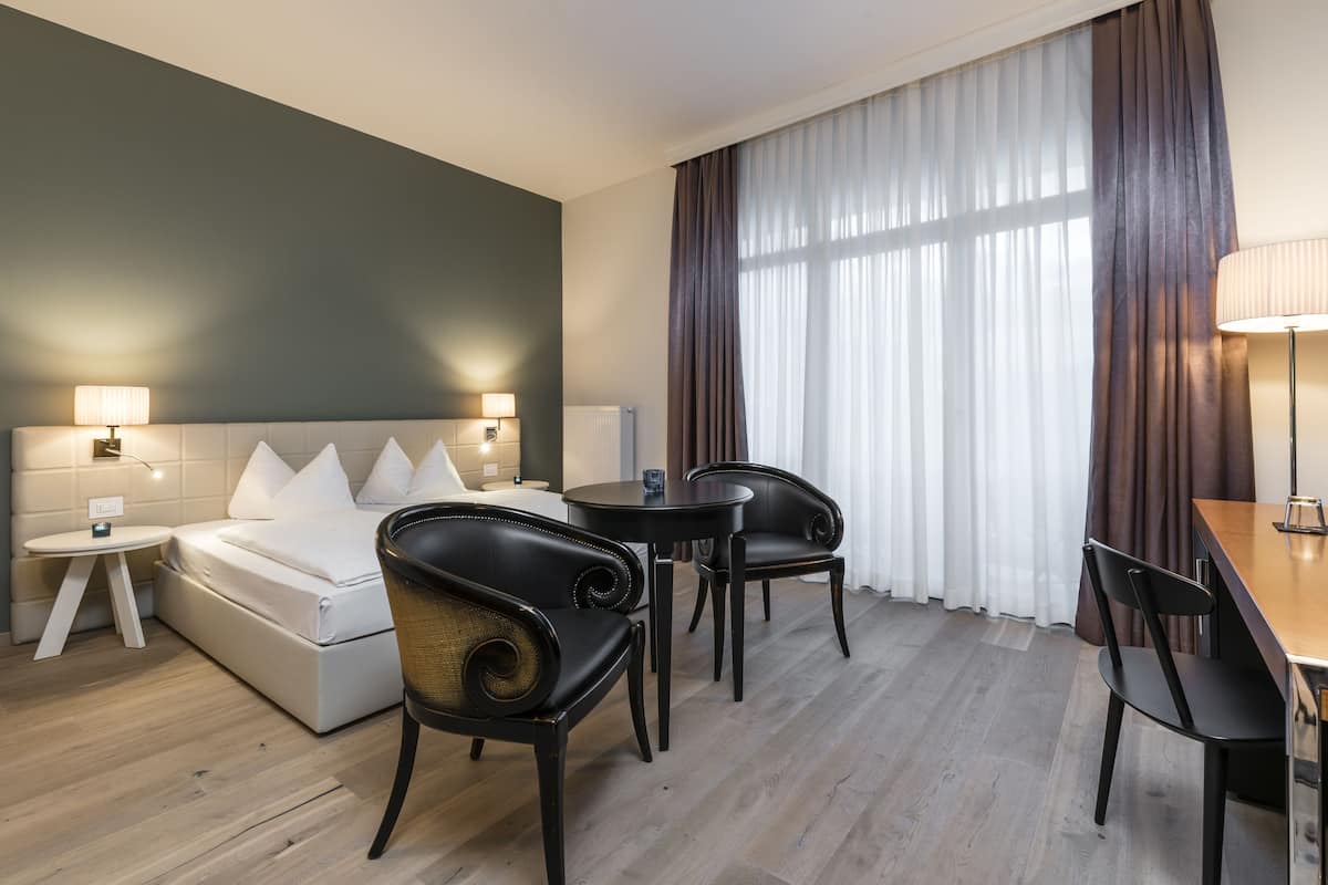 Une chambre d’hôtel avec un lit, un bureau, deux chaises, une lampe et une fenêtre avec des rideaux.