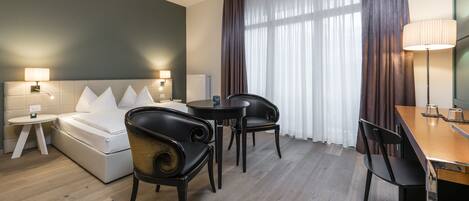 A hotel room with a bed, a desk, two chairs, a lamp, and a window with curtains.