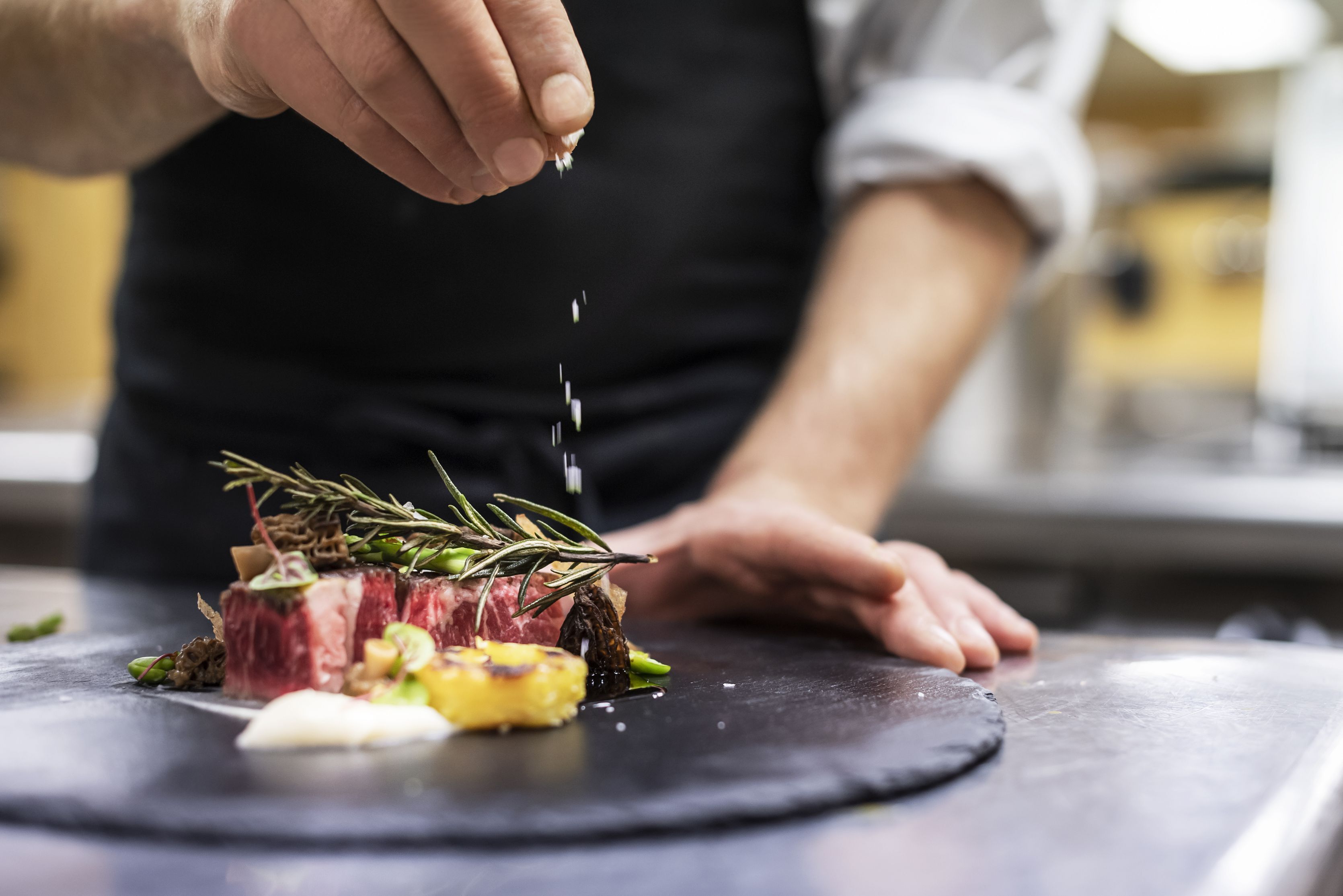 A chef preparing a dish with steak, vegetables, and herbs on a black plate.