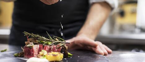 A chef preparing a dish with steak, vegetables, and herbs on a black plate.
