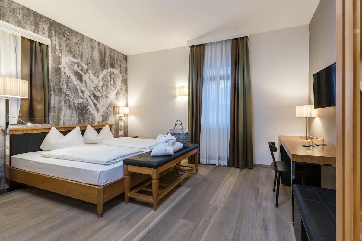 Une chambre d’hôtel avec un lit en bois, un bureau, une chaise, une télévision et une grande fenêtre avec des rideaux.