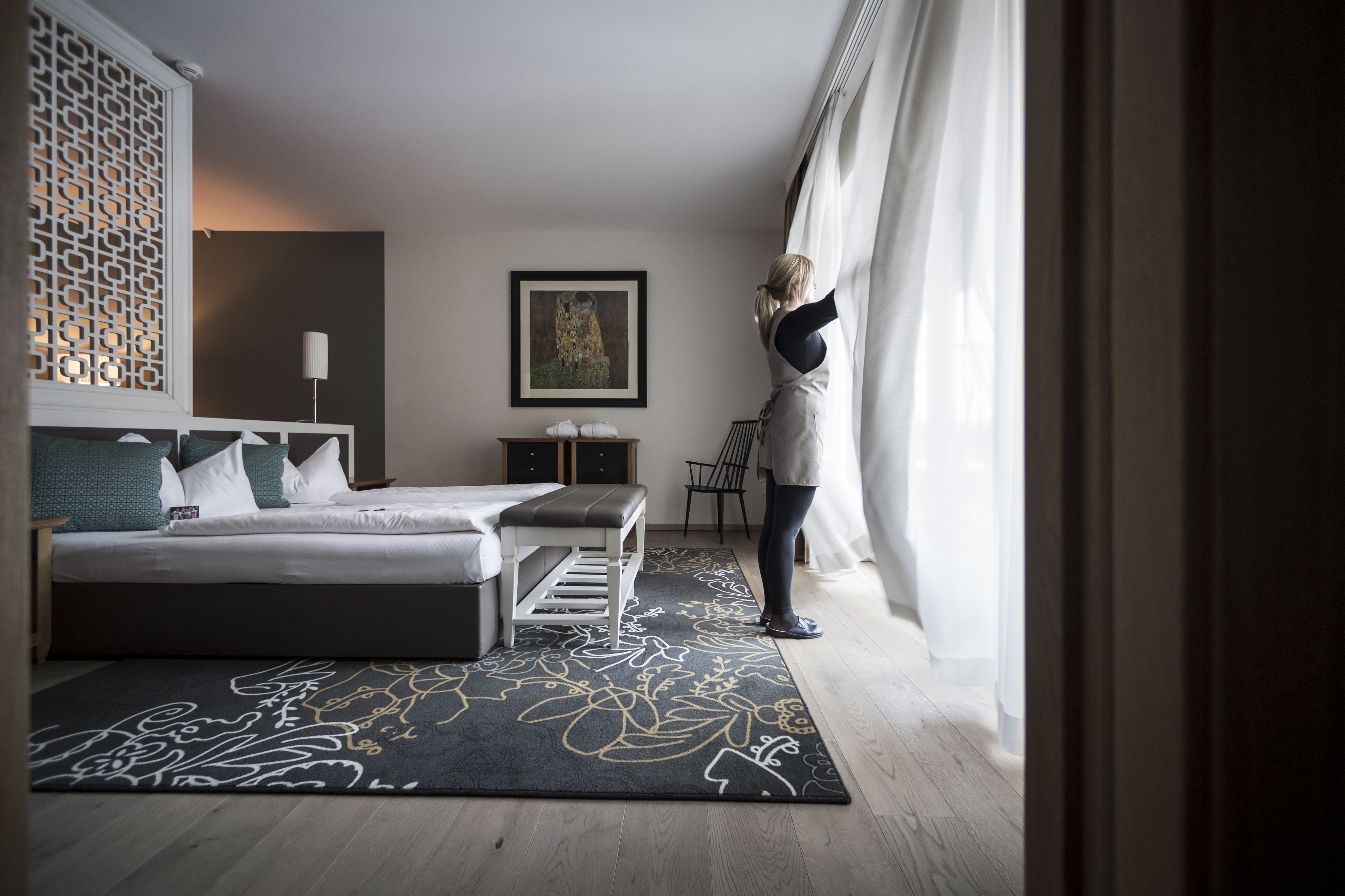 A modern bedroom with a large bed, a bench, a desk, and a chair. There is a decorative wall panel and a framed picture on the wall.