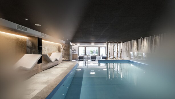 An indoor swimming pool with a modern design, including a seating area with chairs and a table, and a view of the outdoors through large windows. - Post Hotel - Adults Only (San Candido)