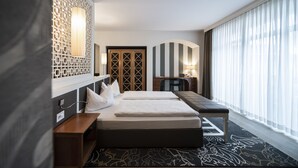 A hotel room with a large bed, a desk, and a computer. There is a window with curtains, a decorative wall panel, and a patterned rug. - Post Hotel - Adults Only (San Candido)