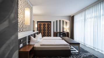 Quarto de hotel com uma cama grande, uma escrivaninha e um computador. Há uma janela com cortinas, um painel decorativo na parede e um tapete estampado.