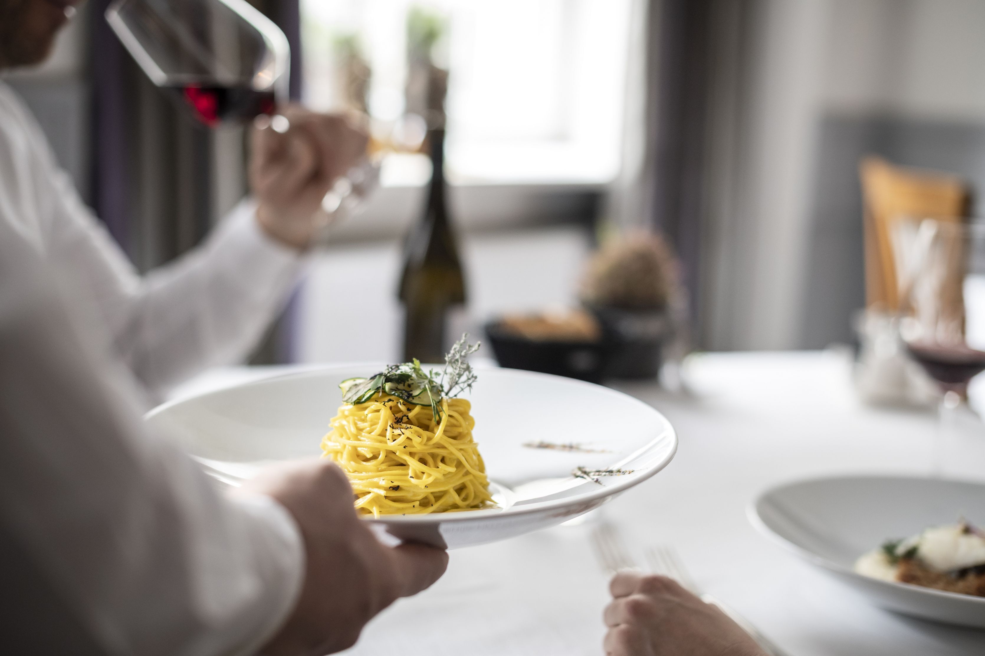A person holding a plate of spaghetti with a garnish, in a dining setting with a bottle of wine and a glass.