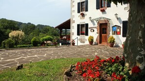 A stone courtyard with a white building featuring a wooden door and shutters, surrounded by greenery and potted plants. - Hotel Rural Irigoienea (Urdax)