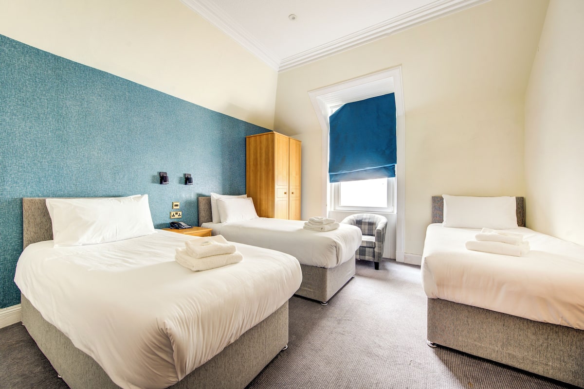 Quarto de hotel com duas camas de solteiro, guarda-roupa de madeira, uma mesinha e uma cadeira.