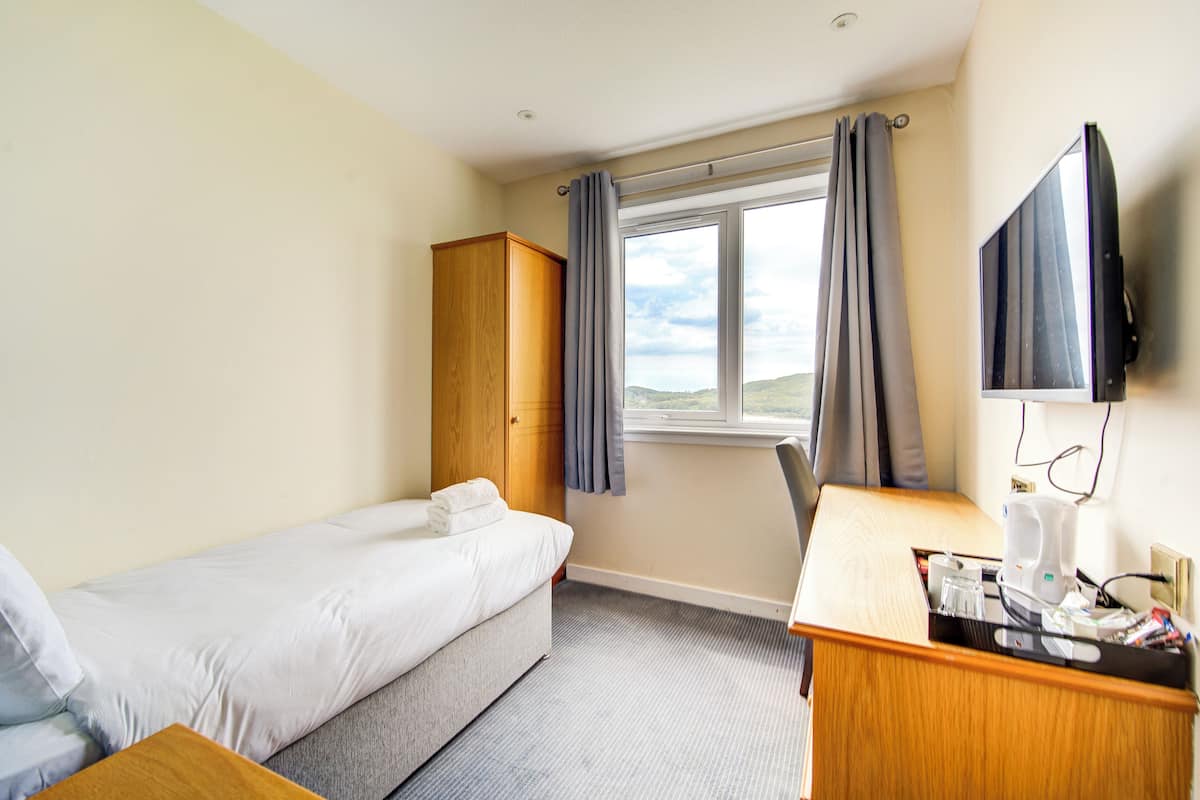 Quarto de hotel com cama, escrivaninha, televisão, janela com cortinas e vista para o exterior.