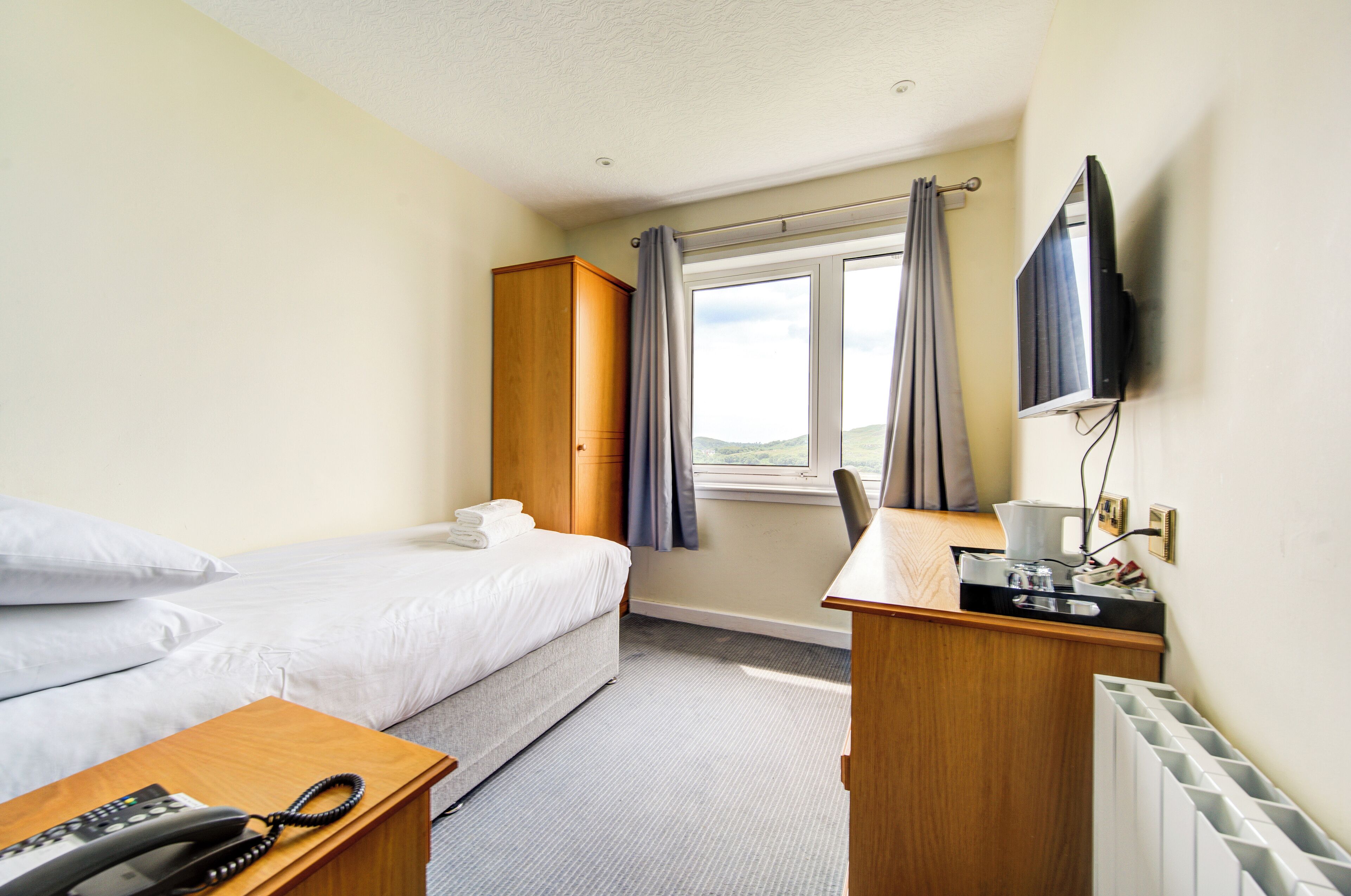 Une chambre d’hôtel avec un lit, un bureau avec un téléphone, une télévision et une fenêtre avec des rideaux.