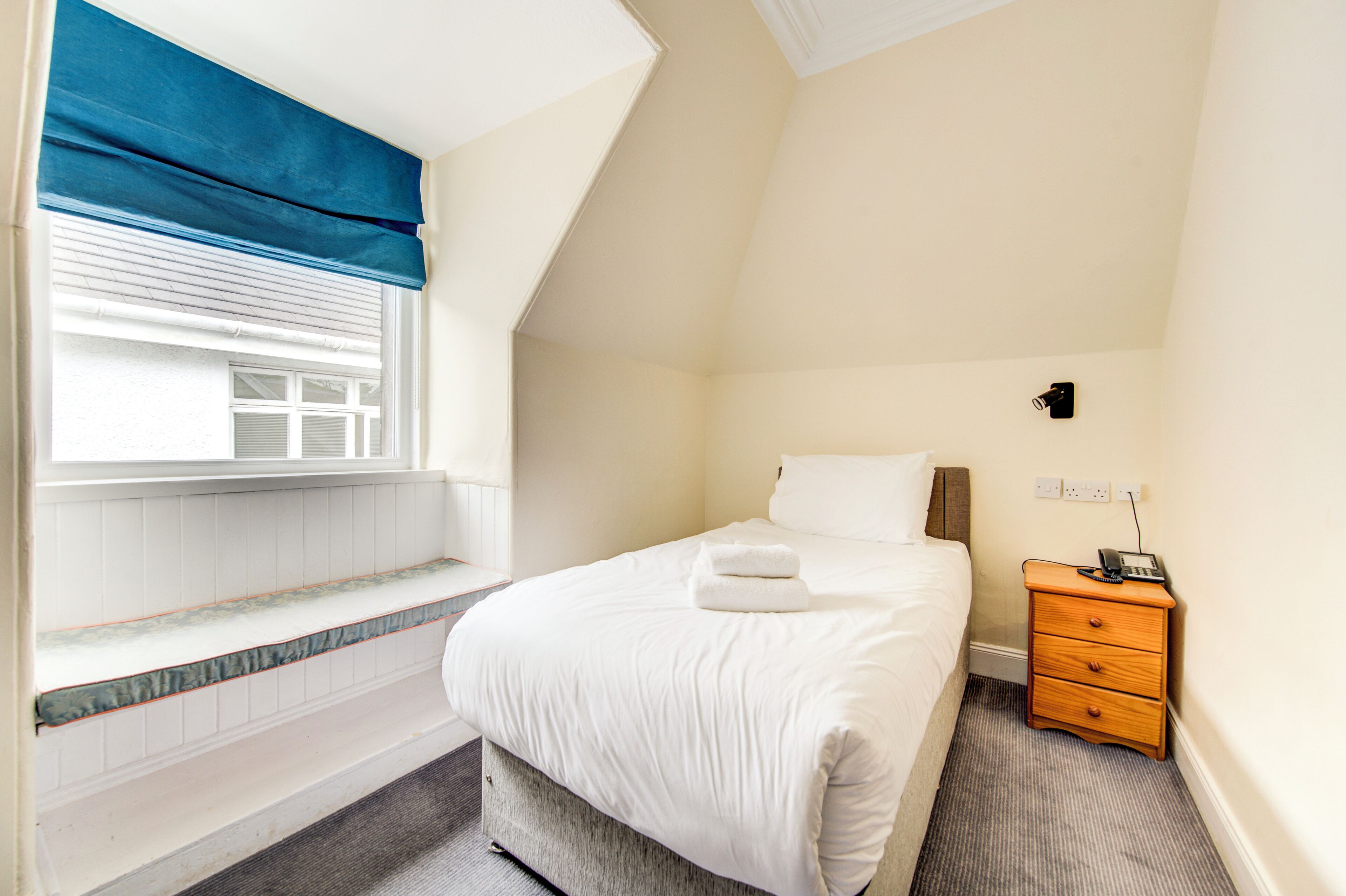 A hotel room with a bed, a bedside table with a phone, a window with blinds, and a small window seat.