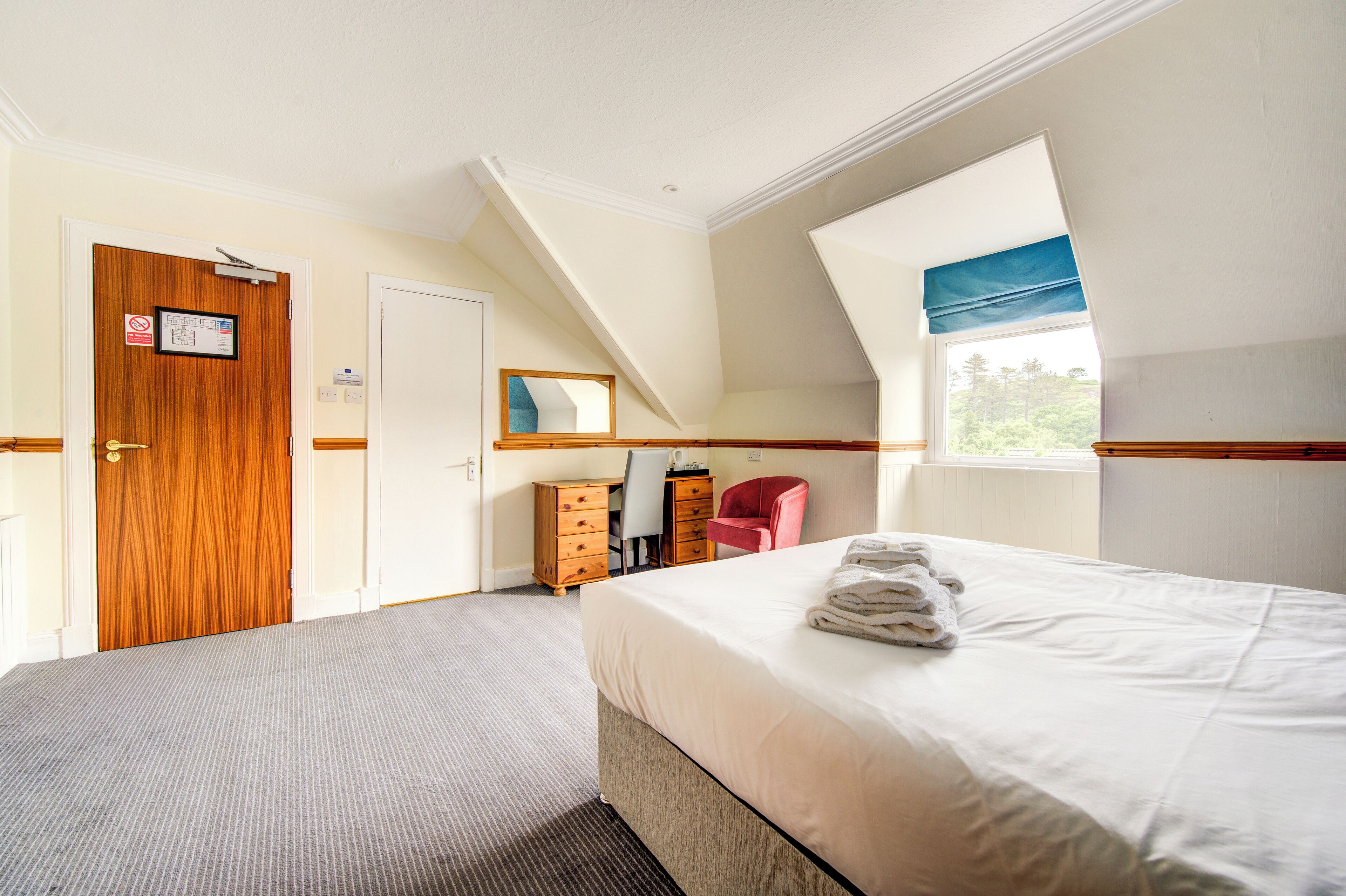 A bedroom with two beds, a wooden door, a desk with a chair, and a window with a view of greenery.