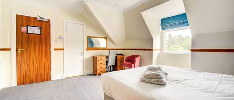 A bedroom with two beds, a wooden door, a desk with a chair, and a window with a view of greenery.