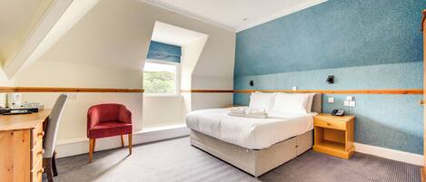 A bedroom with a bed, a desk, a chair, a nightstand, and a window with a view of greenery.