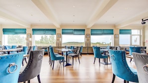 A spacious dining area with blue upholstered chairs and tables, large windows with blue valances, and a view of a distant landscape. - Morar Hotel (Mallaig)