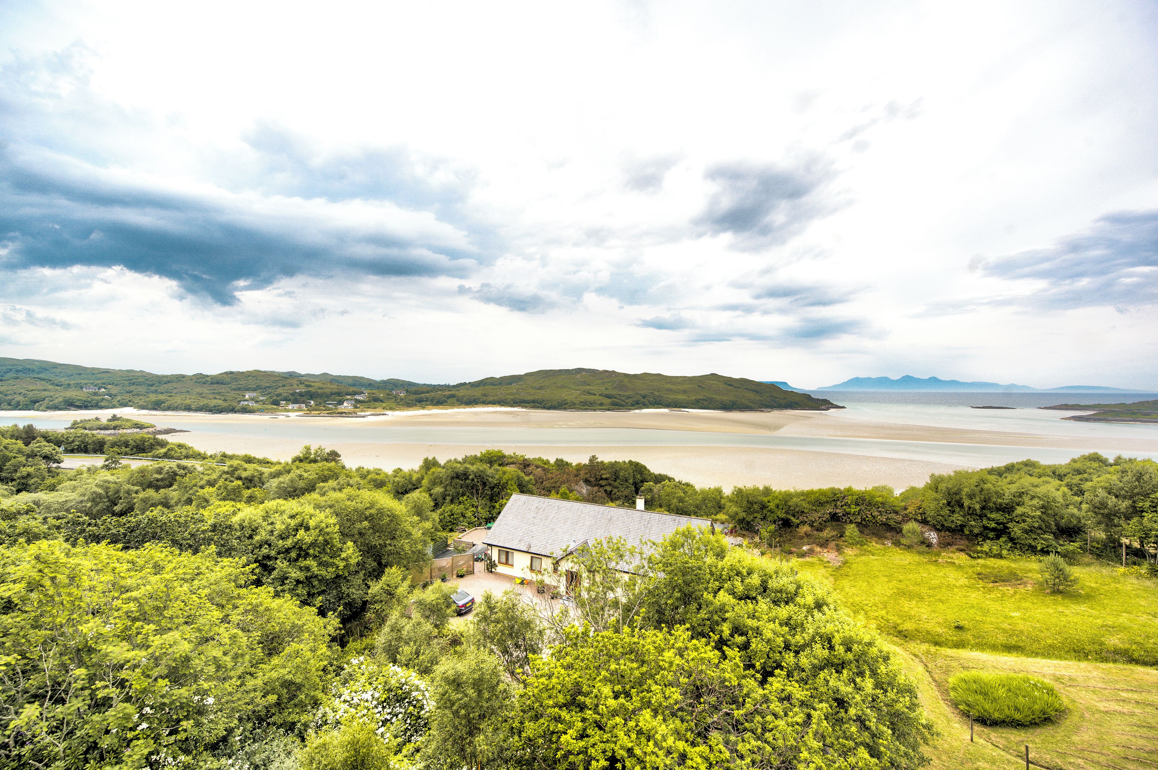 A coastal landscape with a house surrounded by lush greenery and a beach in the background.