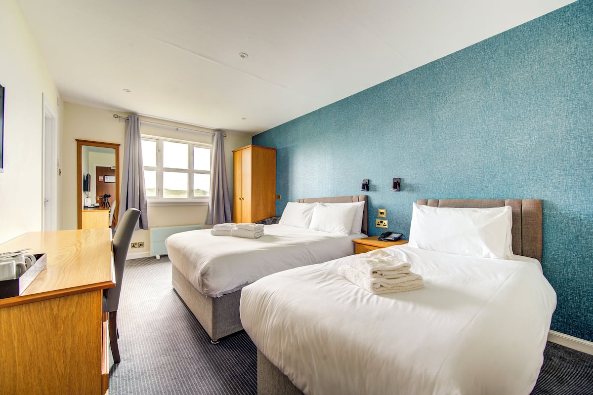 Quarto de hotel com duas camas, uma escrivaninha, uma cadeira e uma janela com cortinas.