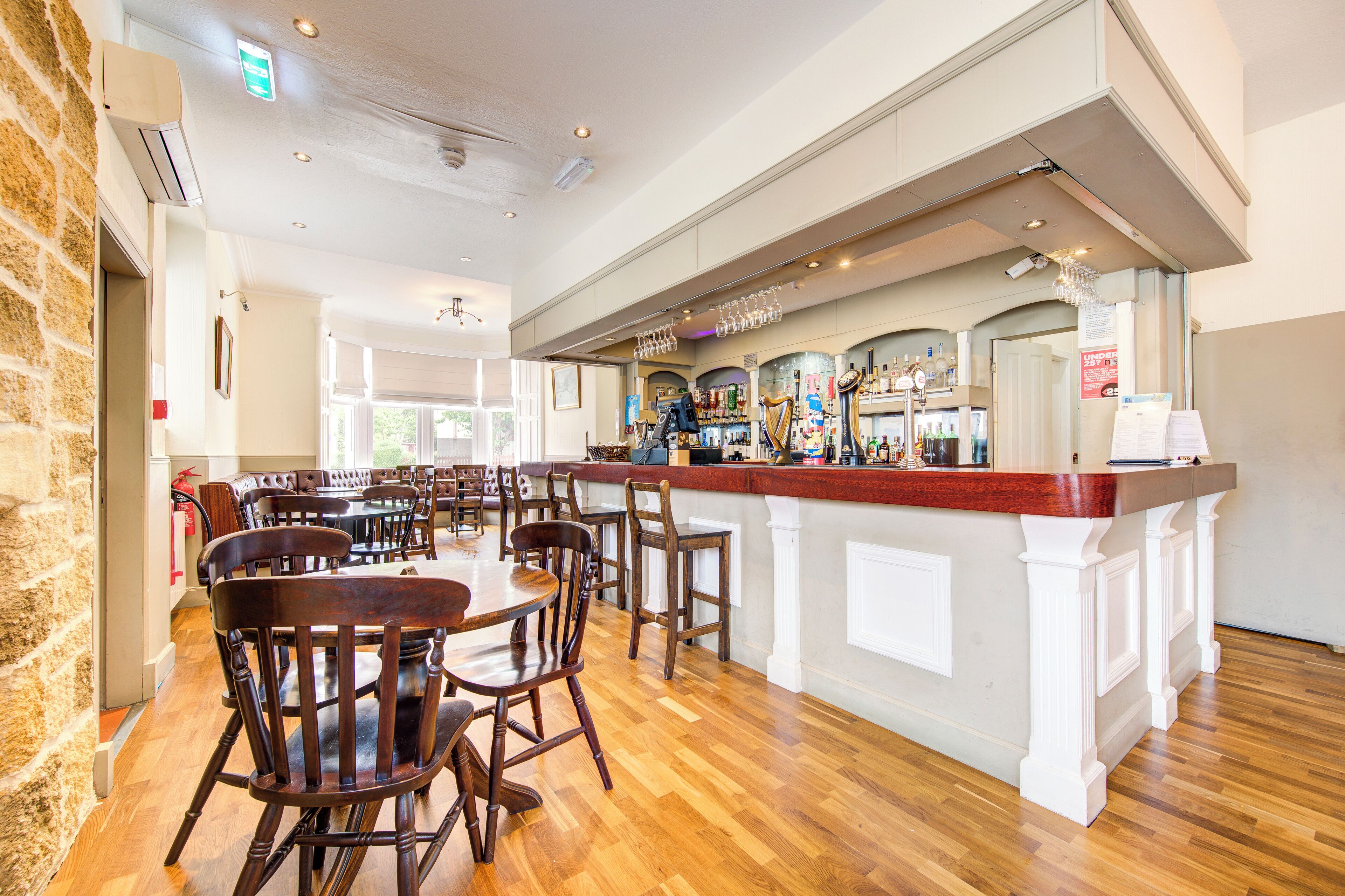 A restaurant with wooden tables and chairs, a bar area with bottles and glasses, and a stone wall on one side.