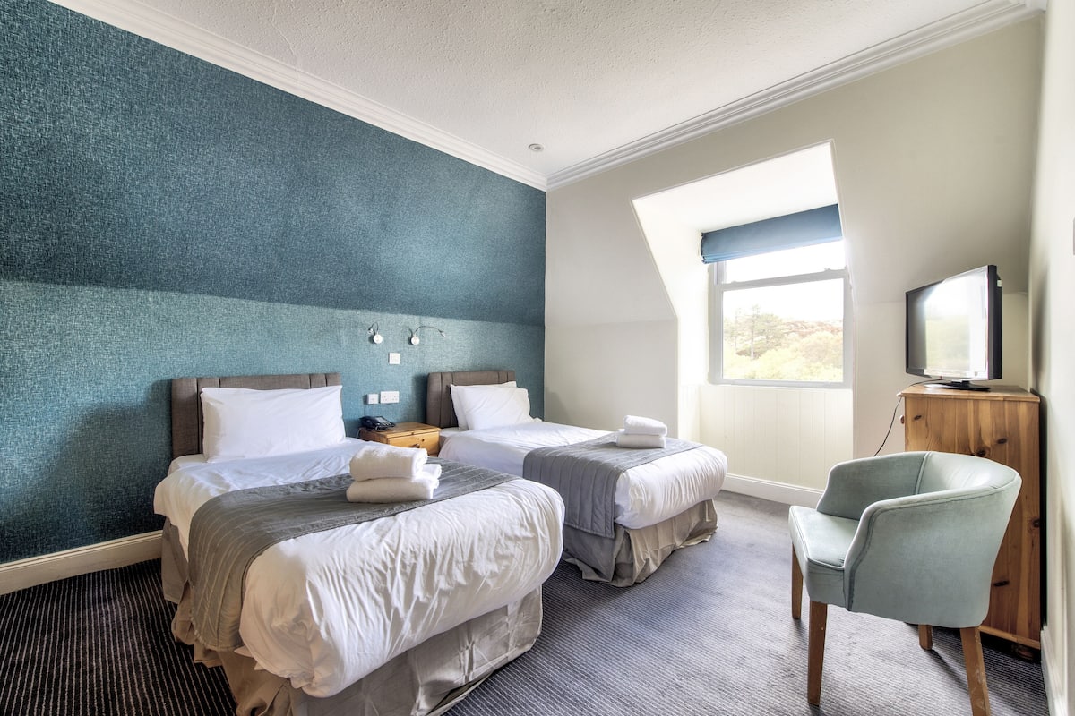 Quarto de hotel com duas camas, televisão, uma cadeira e janela com vista.