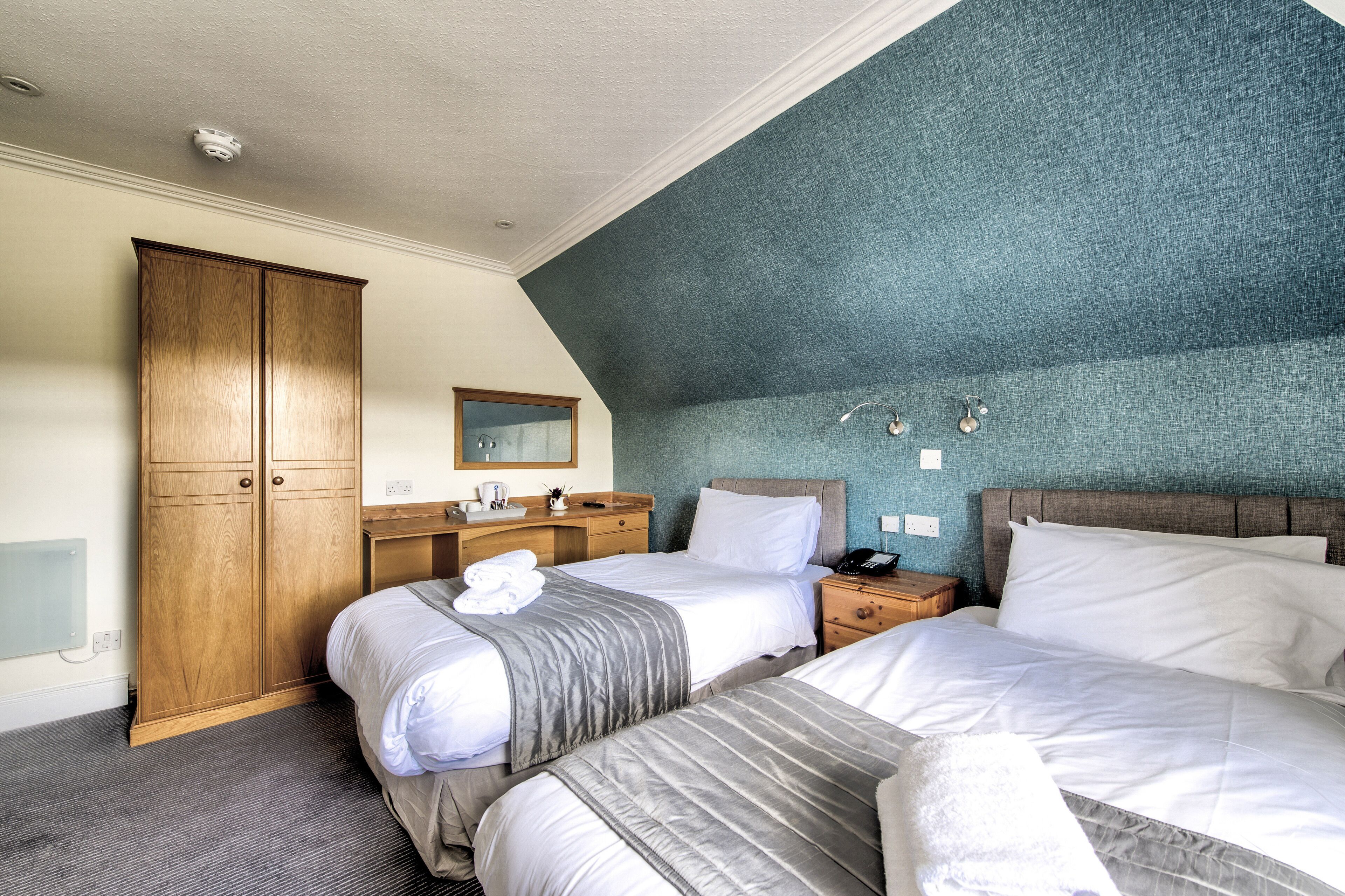 A hotel room with two beds, a wooden wardrobe, a vanity with a mirror, and a blue textured wall.