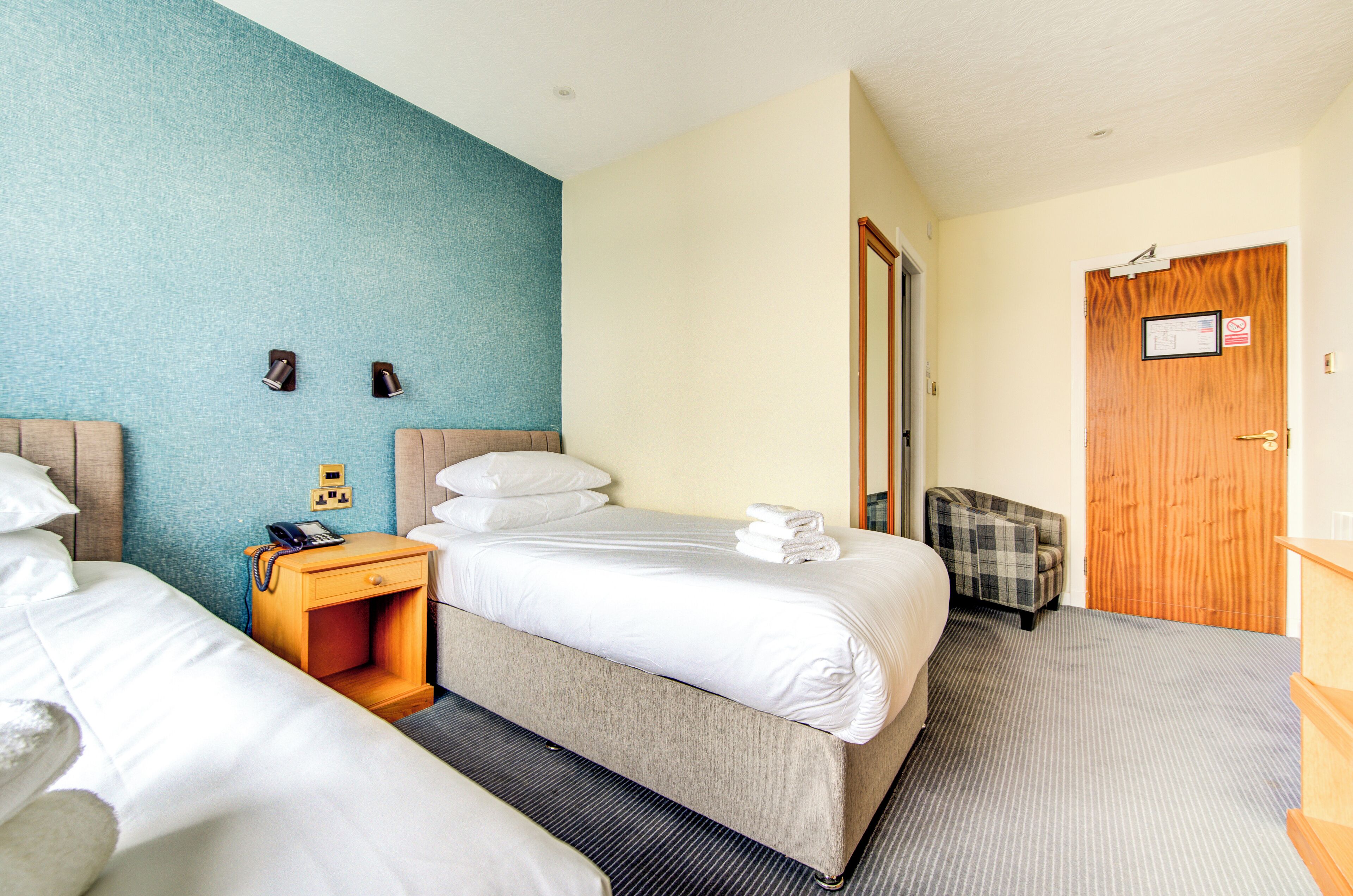 A hotel room with two beds, a wooden nightstand with a phone, a plaid armchair, and a wooden door with a sign.