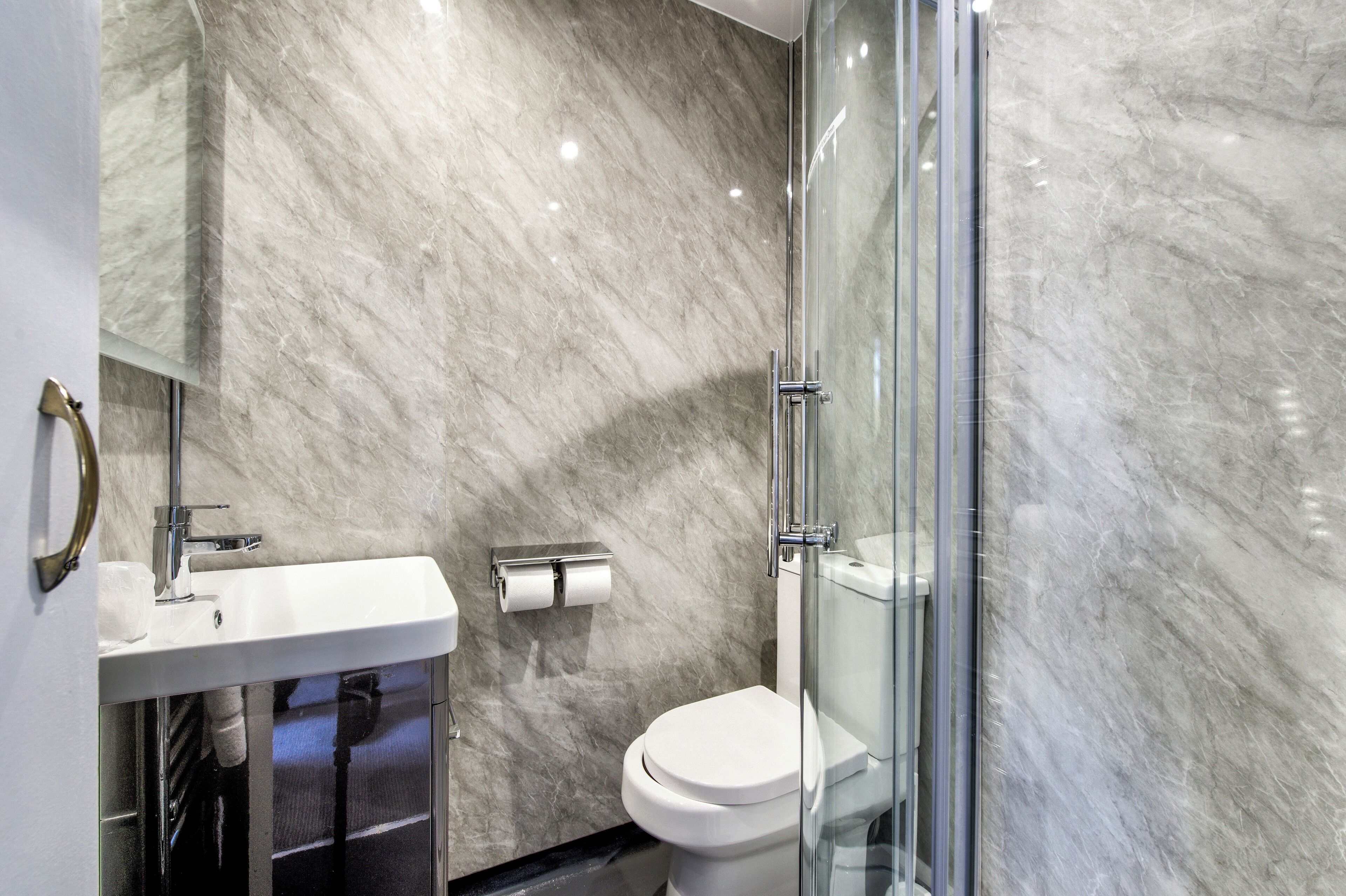 A modern bathroom with a marble wall, a white sink, a glass shower enclosure, and a toilet.