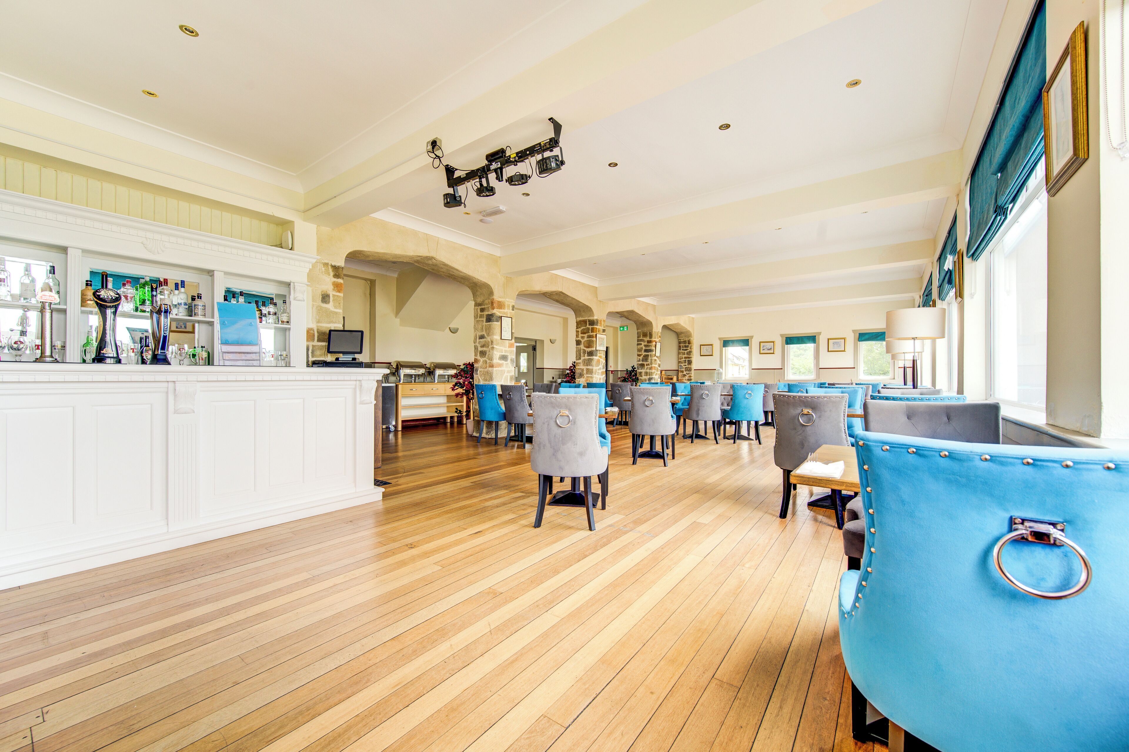 Un hall d’hôtel spacieux doté d’un plancher en bois, de fauteuils recouverts d’un tissu bleu et gris, d’un bar avec diverses bouteilles exposées et d’arcs en pierre.