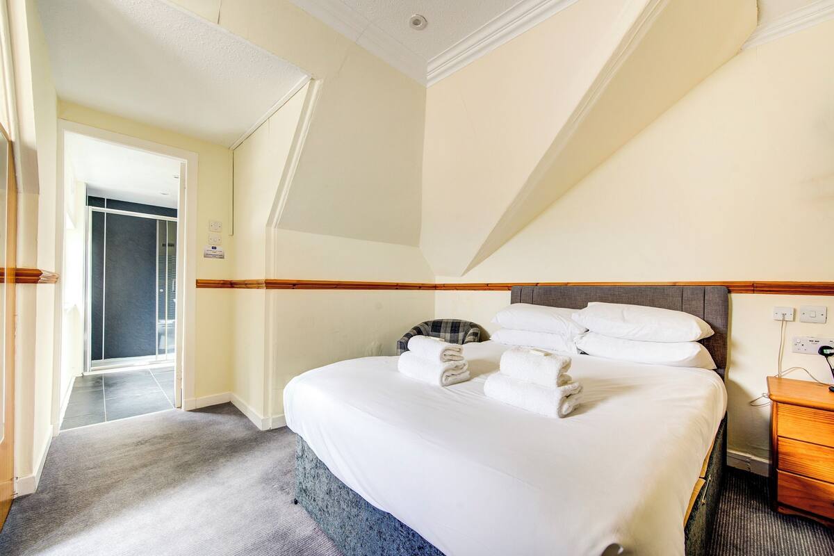 Quarto de hotel com uma cama, uma mesinha de cabeceira de madeira, uma porta que dá para outro quarto e um teto com luminária.