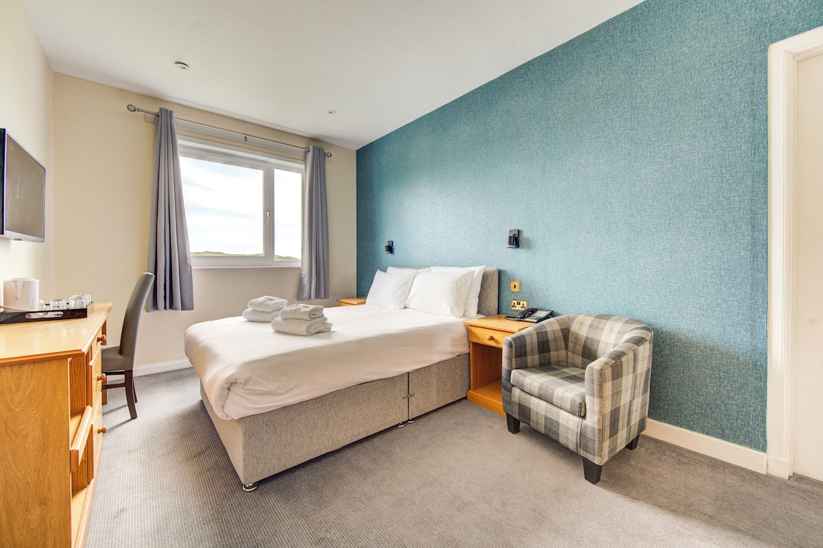 Quarto de hotel com cama, escrivaninha, cadeira e televisão.