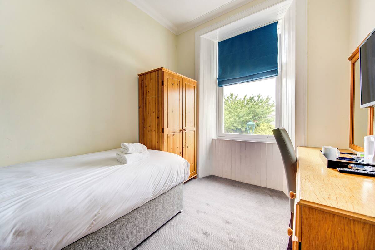 Quarto com uma cama de solteiro, guarda-roupa de madeira, escrivaninha com cadeira e janela com vista para o verde.