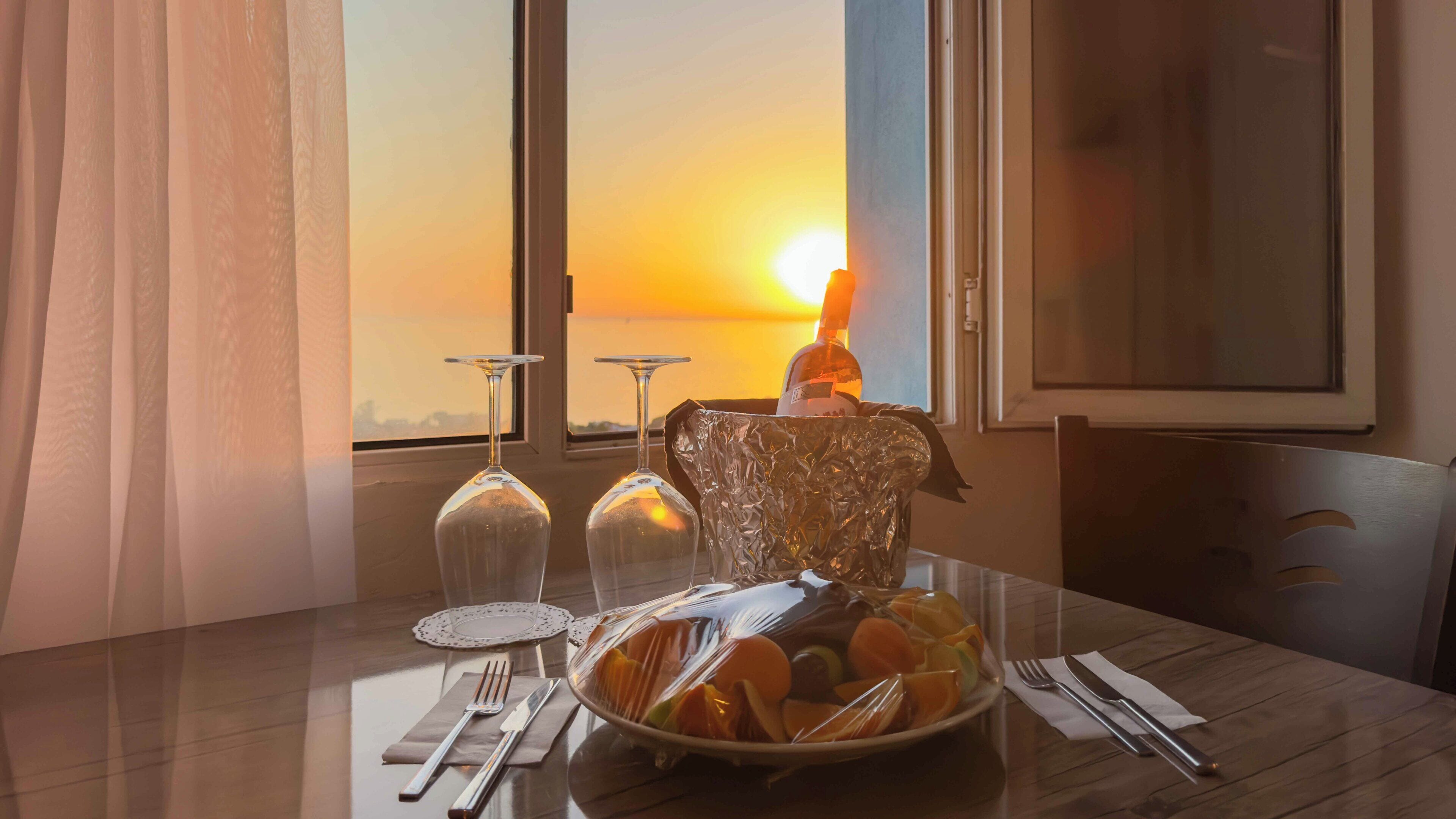 A dining table with a fruit bowl, wine glasses, and cutlery, set against a sunset view through a window.